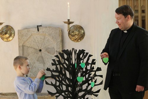 Pappi ja pieni poika ripustavat kasteenpuun lehteä Naantalin kirkossa. Kastepuu kuvassa keskellä. Pappi ja poika seisovat molemmin puolin kastepuuta.