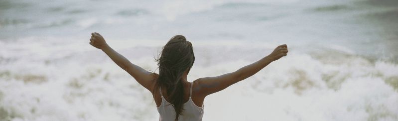 Tyttö seisoo kuohuvan meren äärellä kädet levitettyinä ilmaan.